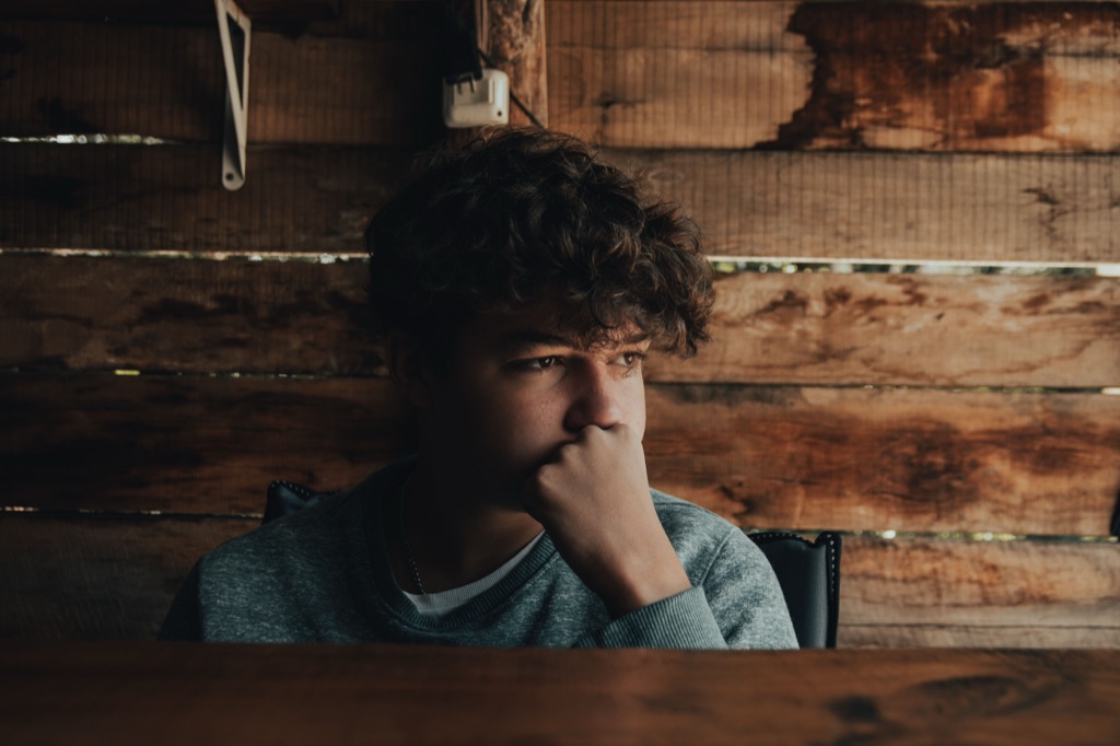 A teenage boy sits at a table, resting his chin on his hand, deep in thought