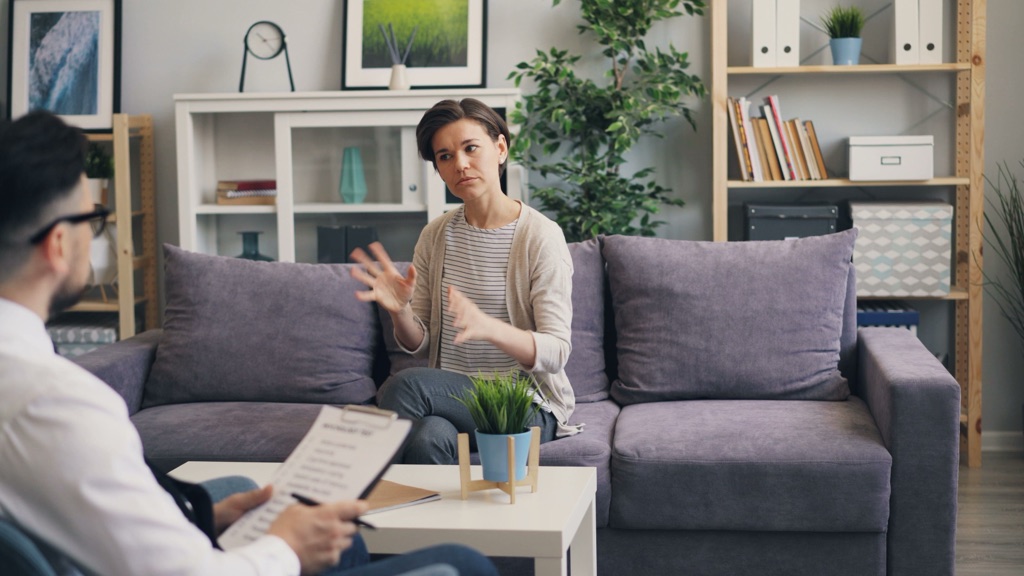 A woman talks with her therapist while sitting on a sofa in a bright counseling office